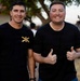 Two men in black t-shirts pose together outdoors, smiling at the camera. The man on the right is giving two thumbs up and wearing a ring. The left man's shirt displays a military-style insignia with crossed cannons and the number 75, suggesting an artillery unit affiliation.