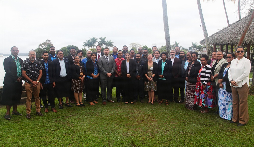 A large group of approximately 30 individuals poses for a formal group photograph outdoors on a grass lawn. The setting appears to be a tropical coastal venue, likely in the Pacific Islands region, with palm trees, a thatched-roof structure, and water visible in the background. The group appears to represent a mix of professional and cultural attire, suggesting an official meeting, conference, or diplomatic event.