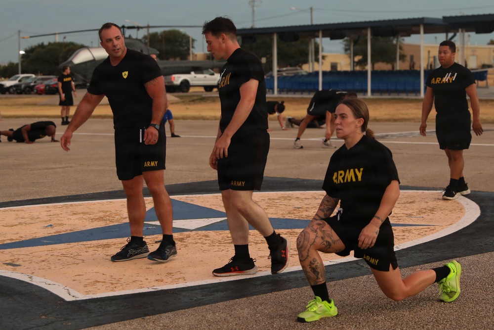A group of U.S. Army personnel are conducting physical training (PT) on an outdoor paved area, likely a military installation. Multiple soldiers wearing matching black Army PT uniforms are engaged in various exercises including lunges and push-ups. The setting appears to be an early morning workout session on what looks like a painted ceremonial or parade ground.
