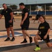 A group of U.S. Army personnel are conducting physical training (PT) on an outdoor paved area, likely a military installation. Multiple soldiers wearing matching black Army PT uniforms are engaged in various exercises including lunges and push-ups. The setting appears to be an early morning workout session on what looks like a painted ceremonial or parade ground.