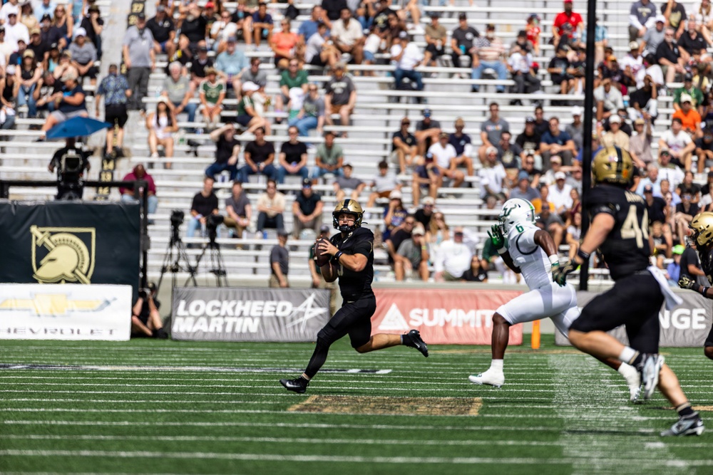 A college football game is in progress, with a quarterback in black uniform preparing to throw a pass while being pursued by a defender in white and green. The home team appears to be Army (West Point), identifiable by the knight/Spartan helmet logo on the sideline banner and black uniforms. The game is being played in a well-attended stadium with sponsors including Lockheed Martin and Chevrolet visible on sideline banners.