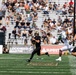 A college football game is in progress, with a quarterback in black uniform preparing to throw a pass while being pursued by a defender in white and green. The home team appears to be Army (West Point), identifiable by the knight/Spartan helmet logo on the sideline banner and black uniforms. The game is being played in a well-attended stadium with sponsors including Lockheed Martin and Chevrolet visible on sideline banners.