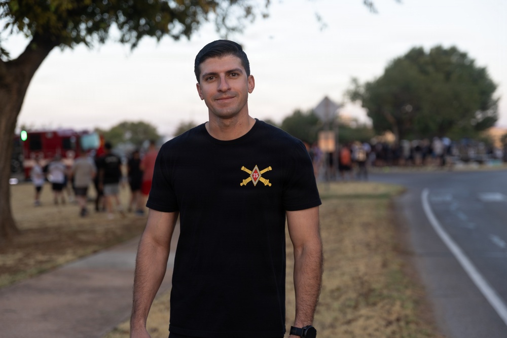 A young man stands on a sidewalk or path near a road, wearing a black t-shirt with a military-style insignia featuring crossed sabers and the number 75. The background shows a blurred outdoor gathering with multiple people, trees, and what appears to be a fire truck on the left side. The setting suggests a community or military event taking place at dusk or early evening.