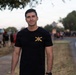 A young man stands on a sidewalk or path near a road, wearing a black t-shirt with a military-style insignia featuring crossed sabers and the number 75. The background shows a blurred outdoor gathering with multiple people, trees, and what appears to be a fire truck on the left side. The setting suggests a community or military event taking place at dusk or early evening.