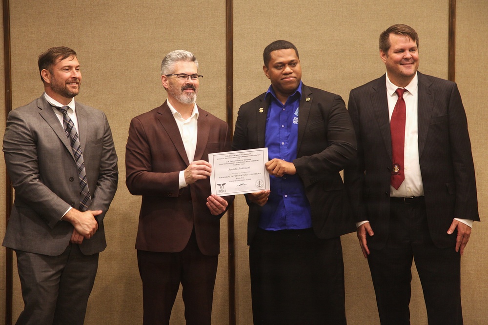 Four men are posing for a formal photograph during what appears to be a certificate presentation ceremony. A man in a blue shirt is jointly holding a certificate with a man in a burgundy suit, suggesting he is the recipient of an award or completion certificate. The setting appears to be an indoor event venue with neutral-toned walls.