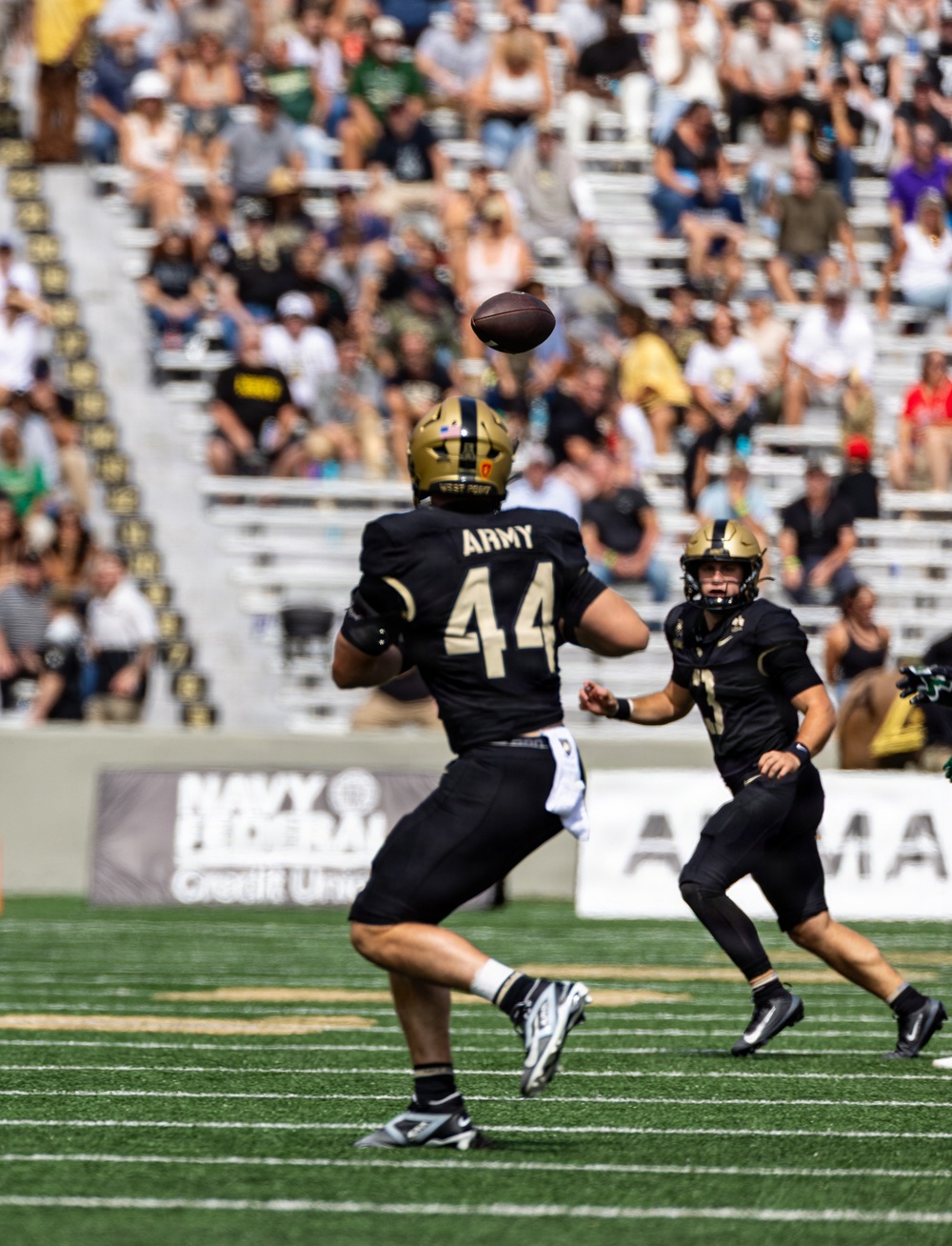 A college football game featuring the Army Black Knights (West Point) is in progress on a green turf field. Player #44 in a black and gold Army jersey is about to catch a pass, with a teammate visible behind him. The stadium is partially filled with spectators watching the daytime game.