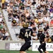A college football game featuring the Army Black Knights (West Point) is in progress on a green turf field. Player #44 in a black and gold Army jersey is about to catch a pass, with a teammate visible behind him. The stadium is partially filled with spectators watching the daytime game.