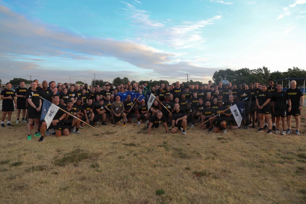 A large group of U.S. Army personnel pose for a group photograph outdoors on a grassy field, likely at a military installation. The majority are wearing black 'ARMY' branded physical training (PT) shirts, suggesting this was taken during or after a physical fitness event. Several unit guidon flags are visible, including ones marked 'IRC' and 'SIS'.