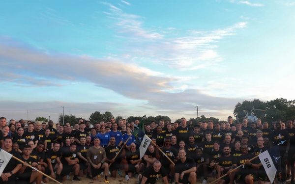 A large group of U.S. Army personnel pose for a group photograph outdoors on a grassy field, likely at a military installation. The majority are wearing black 'ARMY' branded physical training (PT) shirts, suggesting this was taken during or after a physical fitness event. Several unit guidon flags are visible, including ones marked 'IRC' and 'SIS'.