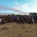 A large group of U.S. Army personnel pose for a group photograph outdoors on a grassy field, likely at a military installation. The majority are wearing black 'ARMY' branded physical training (PT) shirts, suggesting this was taken during or after a physical fitness event. Several unit guidon flags are visible, including ones marked 'IRC' and 'SIS'.