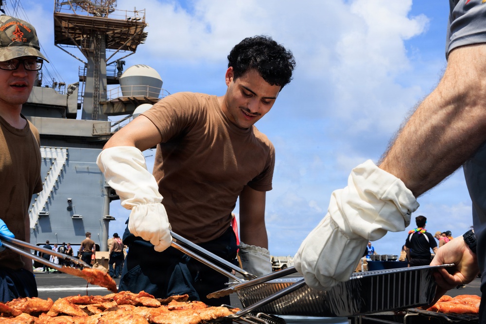 Military personnel are conducting an outdoor barbecue or cookout on the flight deck of a naval vessel. A young sailor wearing a brown t-shirt and white heat-resistant gloves is using tongs to handle grilled meat in aluminum trays. The event appears to be a morale or recreational gathering, with multiple crew members visible in the background against a partly cloudy sky.