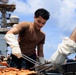Military personnel are conducting an outdoor barbecue or cookout on the flight deck of a naval vessel. A young sailor wearing a brown t-shirt and white heat-resistant gloves is using tongs to handle grilled meat in aluminum trays. The event appears to be a morale or recreational gathering, with multiple crew members visible in the background against a partly cloudy sky.