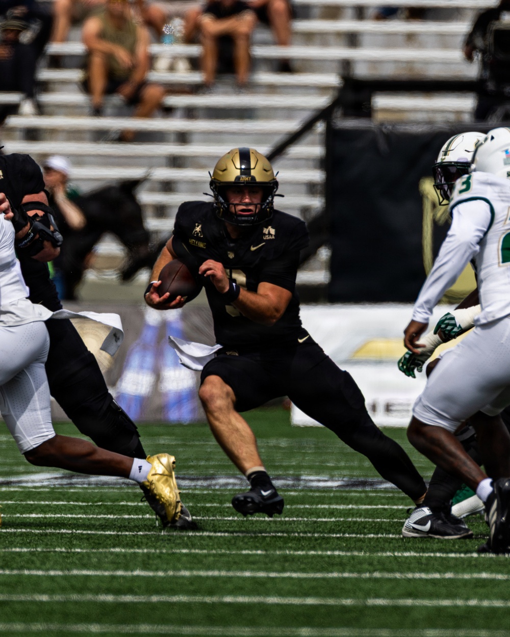 A college football game featuring an Army Black Knights player in black and gold uniform carrying the football through a gap in the defense. The ball carrier is running against opponents wearing white and green uniforms. The action is captured mid-play on a green turf field with stadium seating visible in the background.