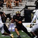 A college football game featuring an Army Black Knights player in black and gold uniform carrying the football through a gap in the defense. The ball carrier is running against opponents wearing white and green uniforms. The action is captured mid-play on a green turf field with stadium seating visible in the background.