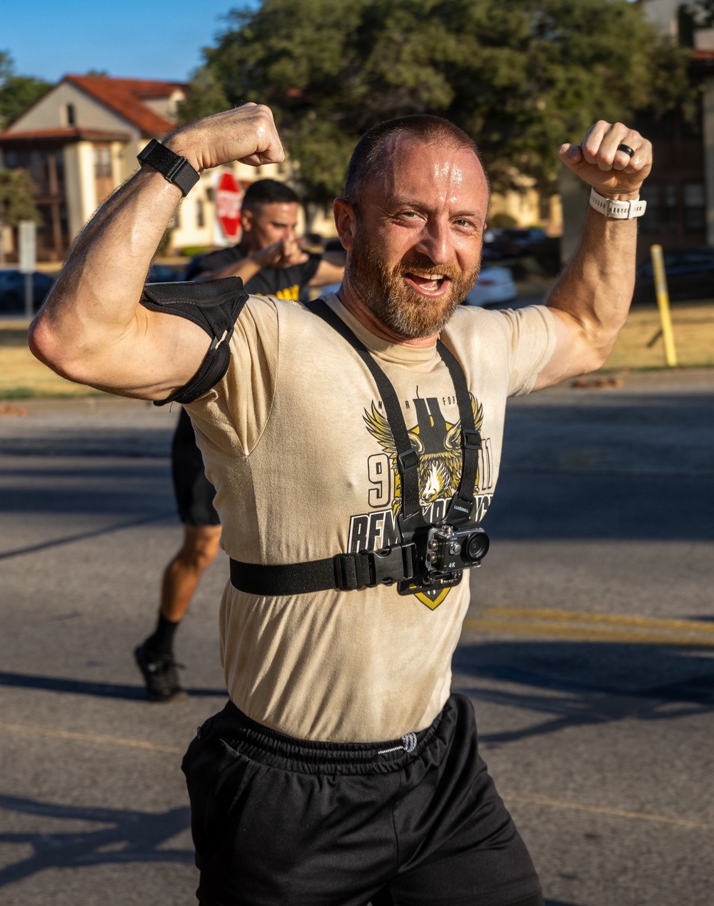 A muscular, bearded man with a shaved head is flexing both arms in a celebratory pose, visibly sweating after physical exertion. He is wearing a tan graphic t-shirt with a chest-mounted action camera (labeled 4K) and black shorts. The setting appears to be an outdoor paved area, likely a military base or institutional campus.