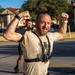 A muscular, bearded man with a shaved head is flexing both arms in a celebratory pose, visibly sweating after physical exertion. He is wearing a tan graphic t-shirt with a chest-mounted action camera (labeled 4K) and black shorts. The setting appears to be an outdoor paved area, likely a military base or institutional campus.