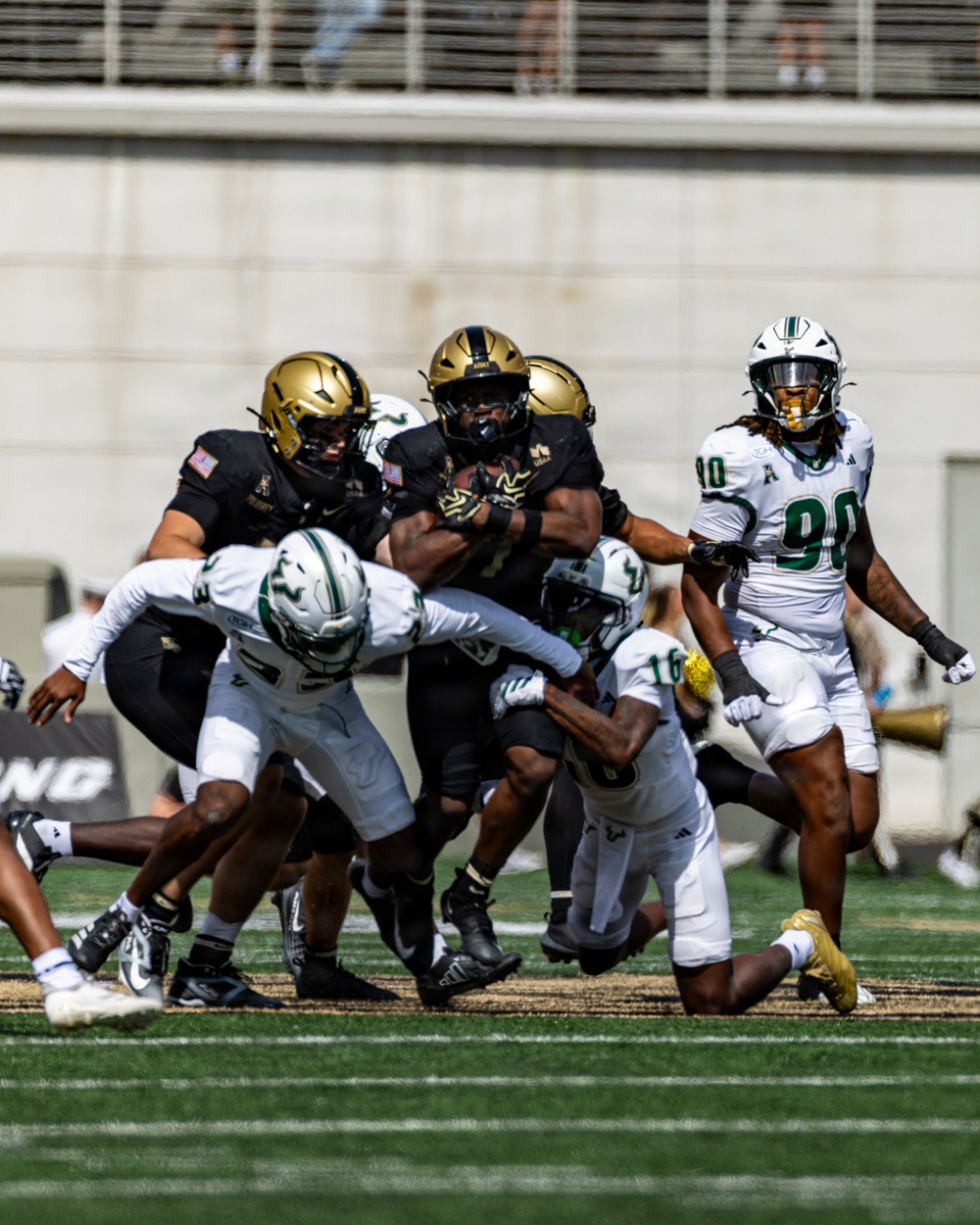 A college football game in progress showing a ball carrier in black and gold uniform being tackled by multiple defenders in white and green uniforms. The offensive team wearing black jerseys with gold helmets appears to be Army (West Point), while the defensive team in white with green accents appears to be South Florida (USF). Multiple players are engaged in the tackle attempt near the line of scrimmage.
