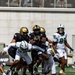 A college football game in progress showing a ball carrier in black and gold uniform being tackled by multiple defenders in white and green uniforms. The offensive team wearing black jerseys with gold helmets appears to be Army (West Point), while the defensive team in white with green accents appears to be South Florida (USF). Multiple players are engaged in the tackle attempt near the line of scrimmage.