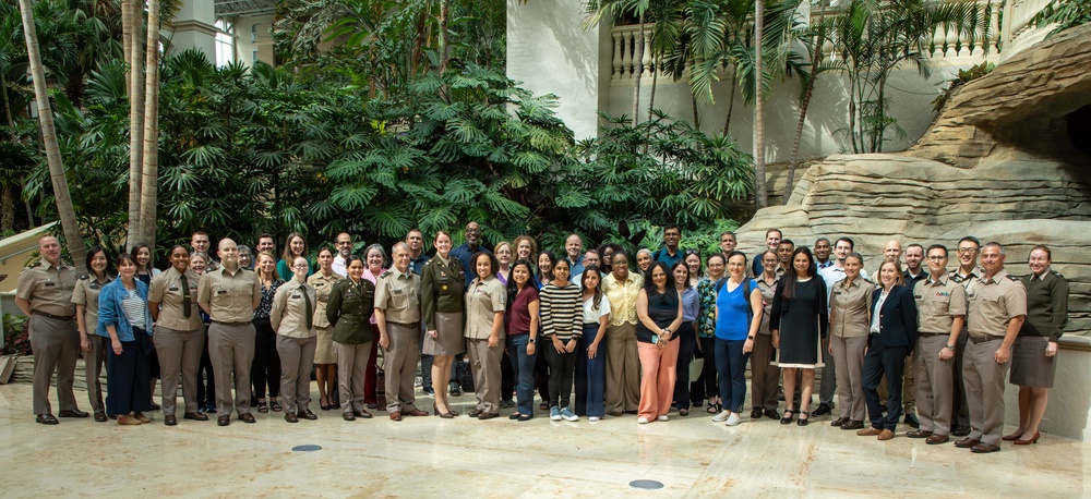 A large group of approximately 50 people pose for a formal group photograph in what appears to be an indoor atrium or conservatory with tropical vegetation. The group consists of a mix of military personnel in tan/khaki uniforms and civilians in business casual attire. The setting features lush palm trees, tropical plants, and a decorative rock formation in the background.