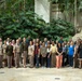 A large group of approximately 50 people pose for a formal group photograph in what appears to be an indoor atrium or conservatory with tropical vegetation. The group consists of a mix of military personnel in tan/khaki uniforms and civilians in business casual attire. The setting features lush palm trees, tropical plants, and a decorative rock formation in the background.