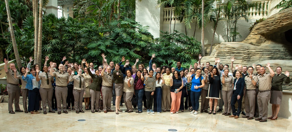 A large group of approximately 50-60 people pose for a formal group photograph in what appears to be an indoor atrium or conservatory with tropical vegetation. Many individuals are wearing tan/khaki uniforms consistent with a government agency or park service, while others are in civilian business casual attire. The group is waving at the camera in a celebratory or welcoming gesture.