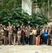 A large group of approximately 50-60 people pose for a formal group photograph in what appears to be an indoor atrium or conservatory with tropical vegetation. Many individuals are wearing tan/khaki uniforms consistent with a government agency or park service, while others are in civilian business casual attire. The group is waving at the camera in a celebratory or welcoming gesture.