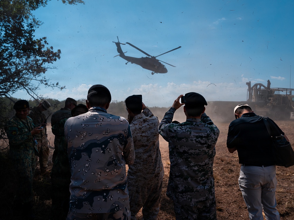 A group of military personnel in various camouflage uniforms observe and photograph a helicopter flying low overhead in a dusty, open field environment. The helicopter appears to be a UH-60 Black Hawk or similar utility helicopter captured in silhouette against a partly cloudy sky. A large military vehicle is visible in the background to the right.