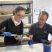 Two researchers wearing blue nitrile gloves are conducting measurements on a small tortoise in what appears to be a field research station or laboratory. The woman on the left is using calipers to measure the tortoise's shell, while the man on the right records data on a printed data sheet. A box labeled 'REPTIBATOR Digital Egg Incubator' is visible in the background, suggesting reptile conservation or research work.