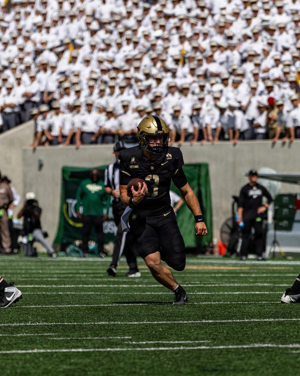 An Army Black Knights football player wearing number 3 in all-black uniform with gold helmet is running with the ball during a game. The stands behind him are filled with what appears to be West Point cadets in white uniforms and caps. The game appears to be played at Michie Stadium, home of Army West Point football.