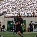 An Army Black Knights football player wearing number 3 in all-black uniform with gold helmet is running with the ball during a game. The stands behind him are filled with what appears to be West Point cadets in white uniforms and caps. The game appears to be played at Michie Stadium, home of Army West Point football.