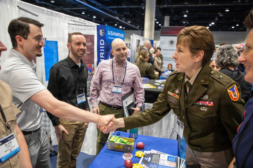 A U.S. Army officer in dress uniform is shaking hands with a civilian attendee at what appears to be a military or defense-related conference expo floor. Several other attendees with conference badges are visible nearby, standing in front of a USAMRIID (U.S. Army Medical Research Institute of Infectious Diseases) booth. The setting is a large convention center with multiple organizational display booths.