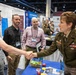 A U.S. Army officer in dress uniform is shaking hands with a civilian attendee at what appears to be a military or defense-related conference expo floor. Several other attendees with conference badges are visible nearby, standing in front of a USAMRIID (U.S. Army Medical Research Institute of Infectious Diseases) booth. The setting is a large convention center with multiple organizational display booths.
