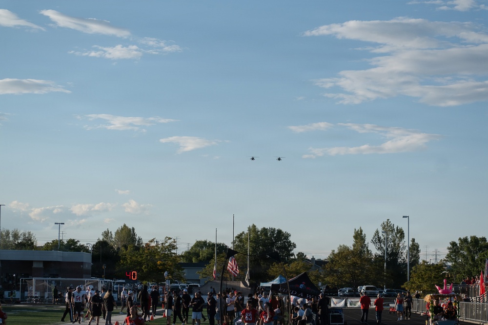 A high school or community football game is taking place on an outdoor field during evening hours with warm golden light. Two aircraft, appearing to be helicopters, are visible flying in formation in the sky above the scene. A crowd of players, coaches, and spectators are gathered along the sidelines near a scoreboard showing '40'.