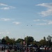 A high school or community football game is taking place on an outdoor field during evening hours with warm golden light. Two aircraft, appearing to be helicopters, are visible flying in formation in the sky above the scene. A crowd of players, coaches, and spectators are gathered along the sidelines near a scoreboard showing '40'.