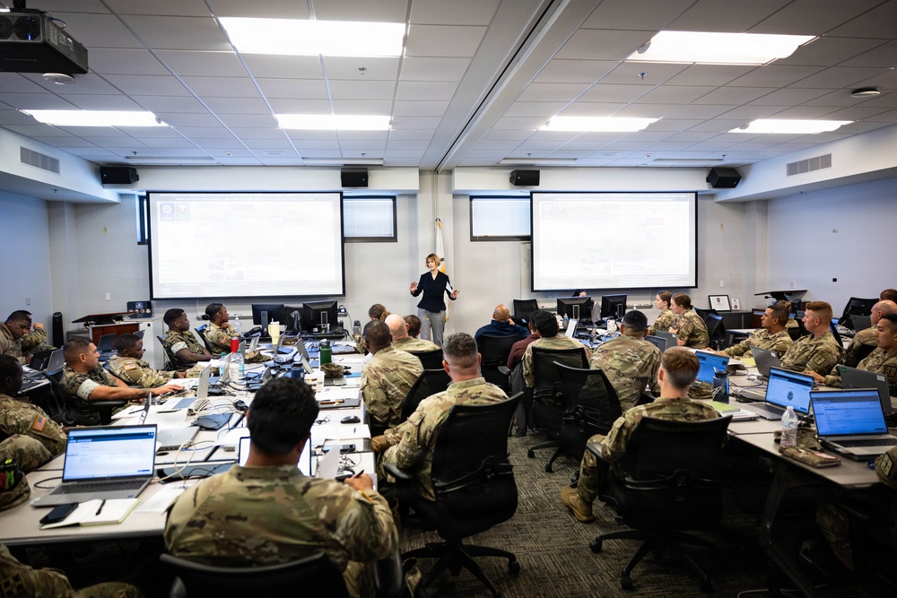 A civilian instructor or presenter stands at the front of a modern classroom briefing a large group of military personnel in camouflage uniforms. The room is equipped with multiple projection screens, laptops, and desktop computers, indicating a technology-focused training or educational session. A flag is visible at the front of the room between the two large projection screens.