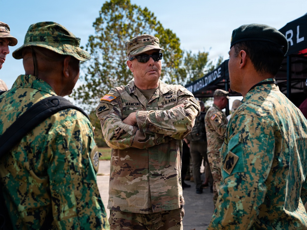 A U.S. Army Major General identified by nameplate as 'MANCINO' stands with arms crossed, engaging in conversation with foreign military personnel wearing green digital camouflage uniforms. The setting appears to be an outdoor military event or exercise, with other uniformed personnel visible in the background. The interaction suggests a multinational military engagement or joint training event.