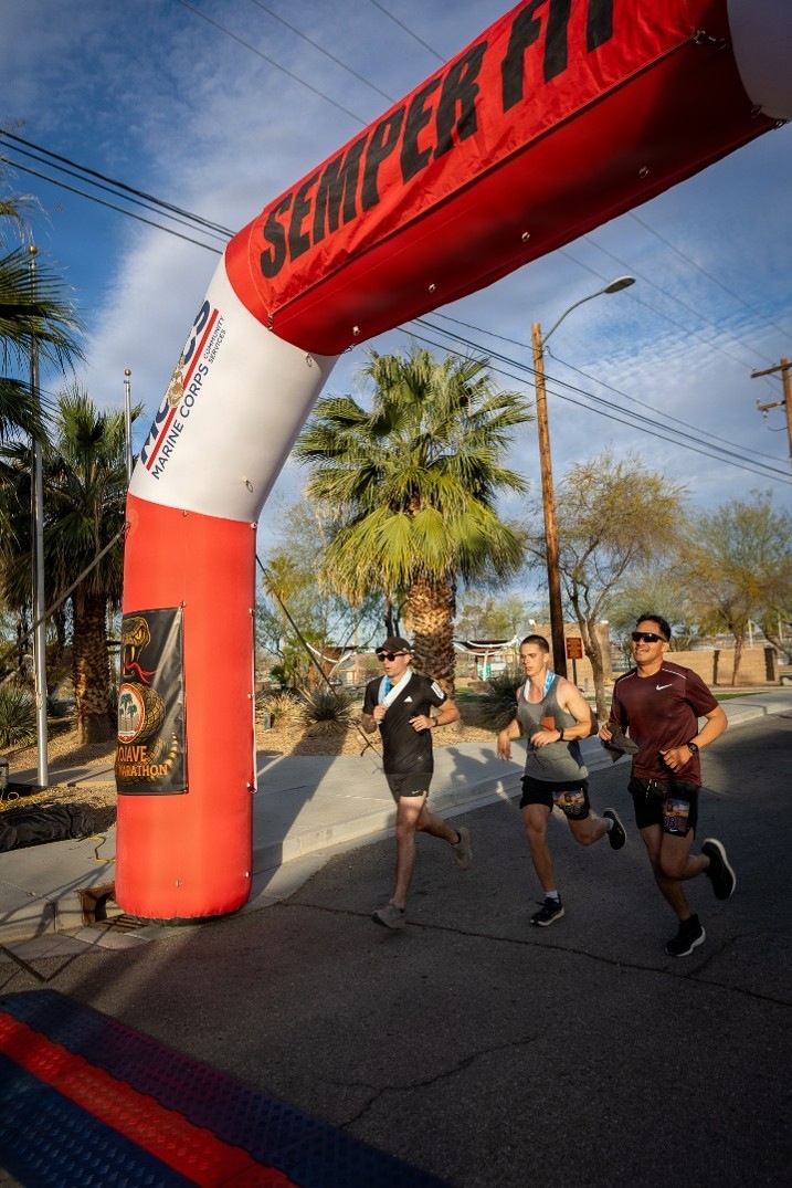 Three runners are crossing or approaching a finish/start line arch at what appears to be the Mojave Marathon, organized under the Marine Corps Community Services 'Semper Fit' program. The event takes place on a paved road in a desert-like environment with palm trees and utility poles visible in the background. Two of the runners are wearing finisher medals, suggesting they have completed the race.