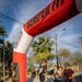 Three runners are crossing or approaching a finish/start line arch at what appears to be the Mojave Marathon, organized under the Marine Corps Community Services 'Semper Fit' program. The event takes place on a paved road in a desert-like environment with palm trees and utility poles visible in the background. Two of the runners are wearing finisher medals, suggesting they have completed the race.