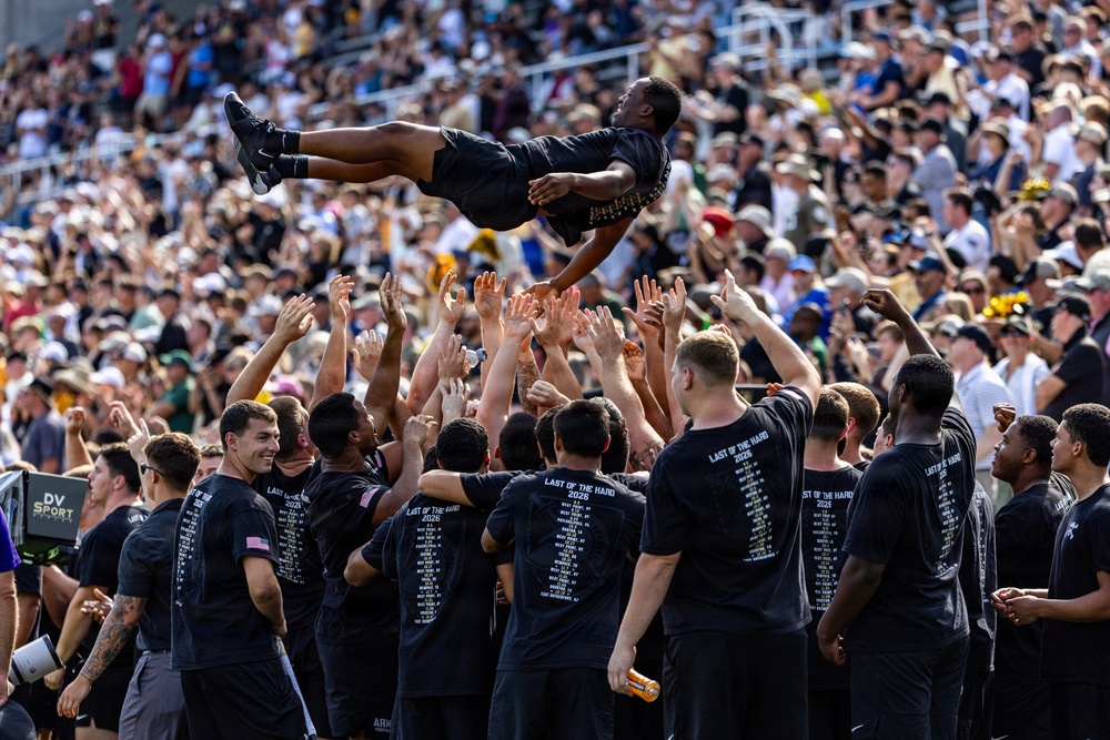 A group of individuals wearing matching black t-shirts reading 'Last of the Hard 2026' are tossing a person high into the air in a celebratory manner at what appears to be a stadium event. The crowd in the bleachers behind them is large and engaged, suggesting this is a significant ceremonial or celebratory occasion. The coordinated matching shirts and the organized group toss indicate a planned team or unit celebration.
