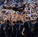 A group of individuals wearing matching black t-shirts reading 'Last of the Hard 2026' are tossing a person high into the air in a celebratory manner at what appears to be a stadium event. The crowd in the bleachers behind them is large and engaged, suggesting this is a significant ceremonial or celebratory occasion. The coordinated matching shirts and the organized group toss indicate a planned team or unit celebration.