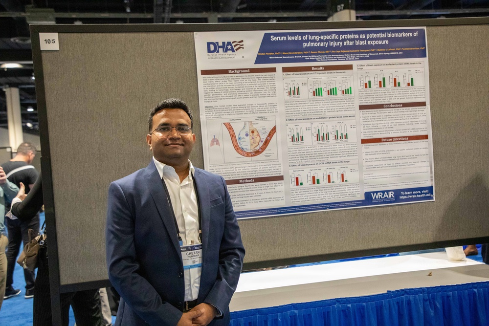 A man named Chetan Pandkar, wearing a blue blazer and conference badge, stands beside a scientific research poster at what appears to be an academic or military medical conference. The poster, sponsored by DHA (Defense Health Agency) and WRAIR, presents research titled 'Serum levels of lung-specific proteins as potential biomarkers of pulmonary injury after blast exposure.' The setting is a large conference hall with other attendees visible in the background.
