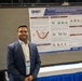 A man named Chetan Pandkar, wearing a blue blazer and conference badge, stands beside a scientific research poster at what appears to be an academic or military medical conference. The poster, sponsored by DHA (Defense Health Agency) and WRAIR, presents research titled 'Serum levels of lung-specific proteins as potential biomarkers of pulmonary injury after blast exposure.' The setting is a large conference hall with other attendees visible in the background.
