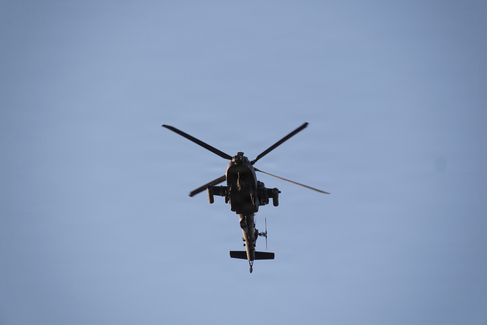 A military attack helicopter is photographed from below against a clear blue sky. The aircraft appears to be in level flight, viewed from a direct underside perspective. The helicopter's silhouette suggests it is a tandem-seat attack variant, consistent with designs like the AH-64 Apache.