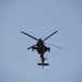 A military attack helicopter is photographed from below against a clear blue sky. The aircraft appears to be in level flight, viewed from a direct underside perspective. The helicopter's silhouette suggests it is a tandem-seat attack variant, consistent with designs like the AH-64 Apache.
