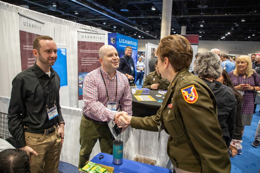 A conference or expo event featuring a USAMRIID (U.S. Army Medical Research Institute of Infectious Diseases) exhibition booth. A civilian man wearing a conference badge is shaking hands with a U.S. Army officer in uniform, while another civilian stands nearby. The venue appears to be a large convention center with multiple booths and attendees visible in the background.