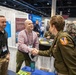 A conference or expo event featuring a USAMRIID (U.S. Army Medical Research Institute of Infectious Diseases) exhibition booth. A civilian man wearing a conference badge is shaking hands with a U.S. Army officer in uniform, while another civilian stands nearby. The venue appears to be a large convention center with multiple booths and attendees visible in the background.