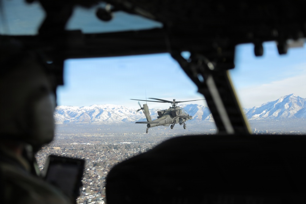 The image is taken from inside a military helicopter cockpit, showing a pilot in the foreground and an attack helicopter (consistent with an AH-64 Apache) flying in formation outside the window. Below is a sprawling urban area, and snow-capped mountain ranges are visible in the background under a clear blue sky. The perspective suggests a formation flight over what appears to be a western U.S. city.