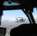 The image is taken from inside a military helicopter cockpit, showing a pilot in the foreground and an attack helicopter (consistent with an AH-64 Apache) flying in formation outside the window. Below is a sprawling urban area, and snow-capped mountain ranges are visible in the background under a clear blue sky. The perspective suggests a formation flight over what appears to be a western U.S. city.