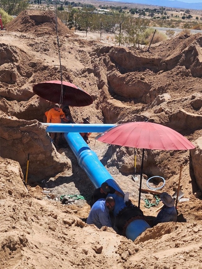 Workers are installing or repairing a large blue water main pipeline in an open excavation trench in what appears to be a desert environment. Multiple workers are positioned in the trench working on pipe connections, shaded by red umbrellas to protect from intense sunlight. The excavation site shows extensive earthwork with sandy, arid soil typical of a southwestern desert region.