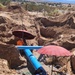 Workers are installing or repairing a large blue water main pipeline in an open excavation trench in what appears to be a desert environment. Multiple workers are positioned in the trench working on pipe connections, shaded by red umbrellas to protect from intense sunlight. The excavation site shows extensive earthwork with sandy, arid soil typical of a southwestern desert region.